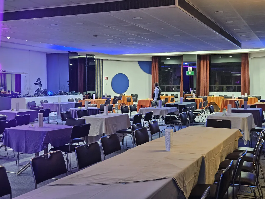 Salón montado: mesa larga vestida en primer plano y mesas redondas con mantel naranja al fondo, bajo luz azul