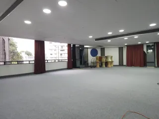 Salón vacío con cortinas rojas, ventanales a la avenida y unos tambores arrumbados a un costado